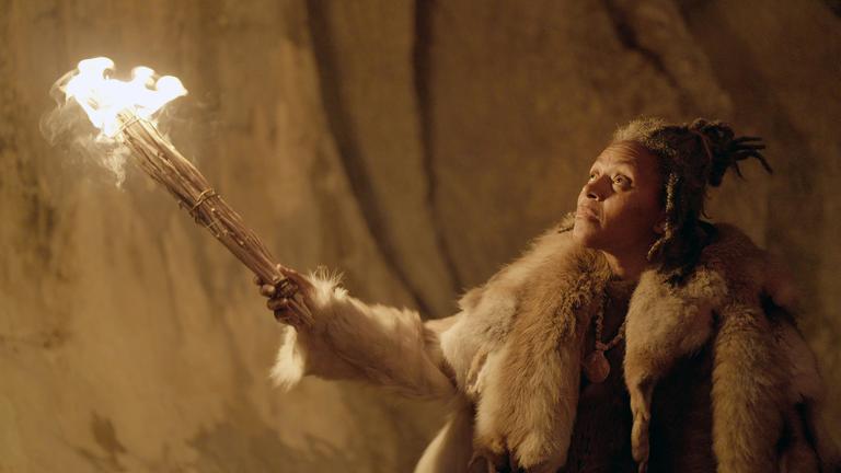 Ein Frau mit einer Fackel leuchtet eine Felswand an. Sie ist in Tierfelle gekleidet und trägt eine Muschel als Halskette.