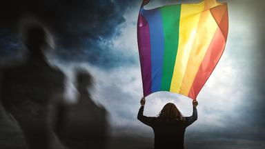 Silhouette einer Person, die mit ausgestreckten Armen eine große Regenbogenfahne über sich hält. Im Hintegrund sind schemenhaft weitere Personen zu erkennen.