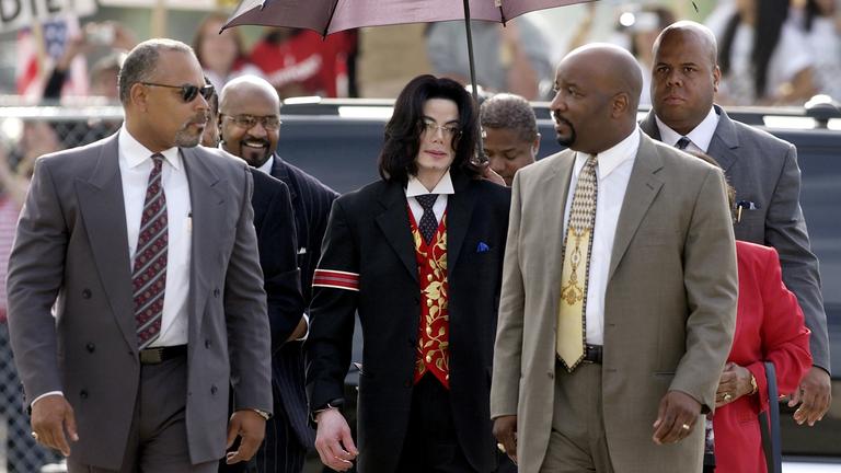 Michael Jackson ist auf dem Weg zum Gericht in Santa Maria im Jahr 2005. Der Popstar ist von seinem Team umgeben. Er trägt einen schwarzen Anzug mit einer auffälligen roten Weste und einer roten Armbinde. Eine Begleiterin hält einen Schirm über ihn.