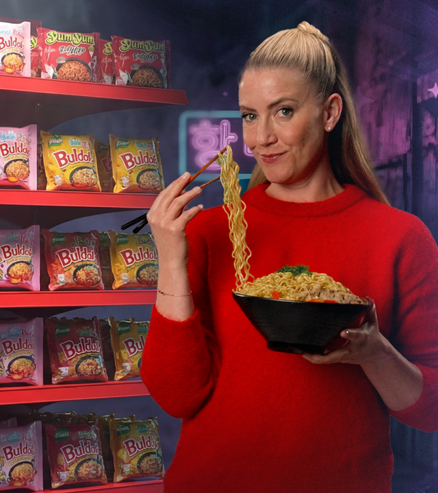 Pia Osterhaus steht mit einer Nudel-Bowl und Essstäbchen in der Han vor einem Regal voller Asia-Tüttensuppen. Mit den Essstäbchen hebt sie Nudeln aus der Bowl Richtung Mund und schaut in die Kamera.