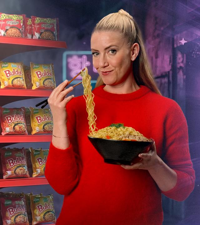 Pia Osterhaus steht mit einer Nudel-Bowl und Essstäbchen in der Han vor einem Regal voller Asia-Tütensuppen. Mit den Essstäbchen hebt sie Nudeln aus der Bowl Richtung Mund und schaut in die Kamera.