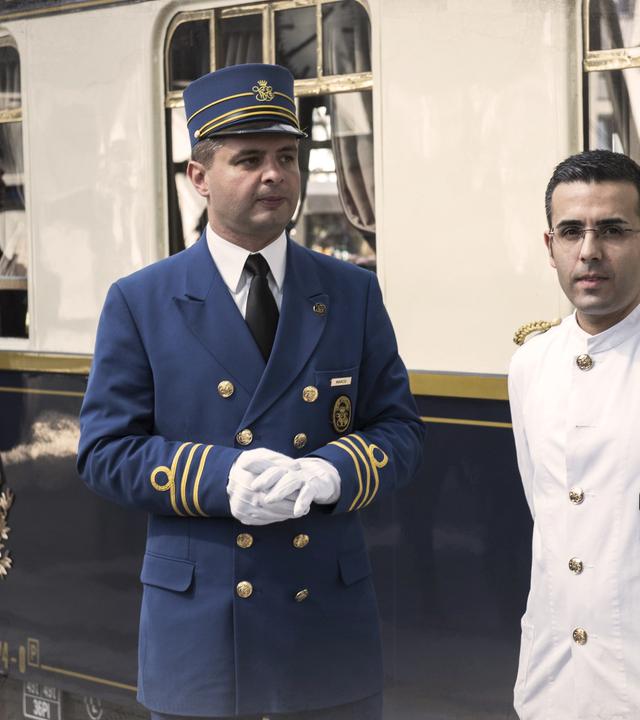 Ein Waggon des Orient-Express an einem Bahnhof, davor zwei Männer vom Zugpersonal. an den Bildrändern leichte Überblendung mit einer alten Landkarte.