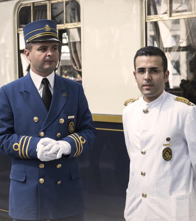 Ein Waggon des Orient-Express an einem Bahnhof, davor zwei Männer vom Zugpersonal. an den Bildrändern leichte Überblendung mit einer alten Landkarte.