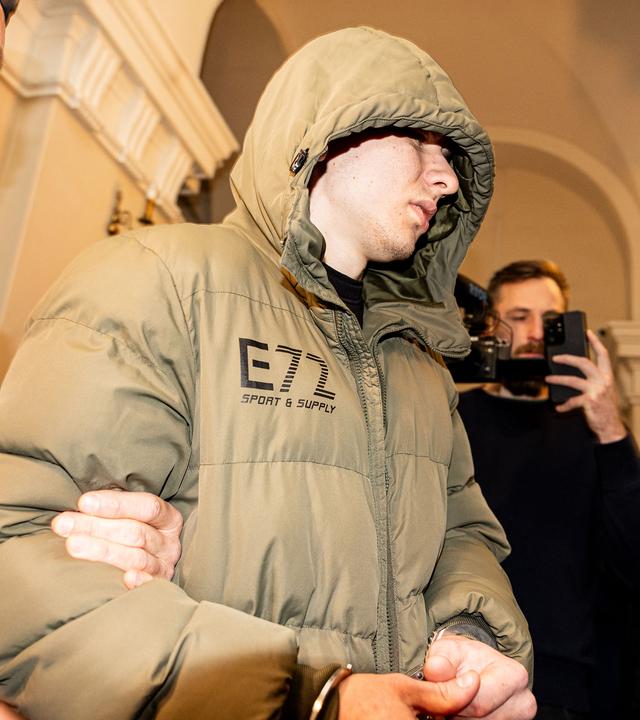 Im Foyer eines klassizistischen Baus wird ein junger Mann in Handschellen und mit olivfarbener Jacke mit Kapuze auf dem Kopf von einem Justizbeamten in schusssicherer Weste geführt.