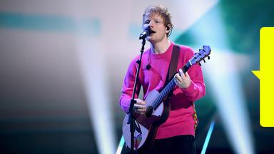 Ed Sheeran singt und spielt Gitarre auf der Bühne. Er trägt einen rosa Pullover.