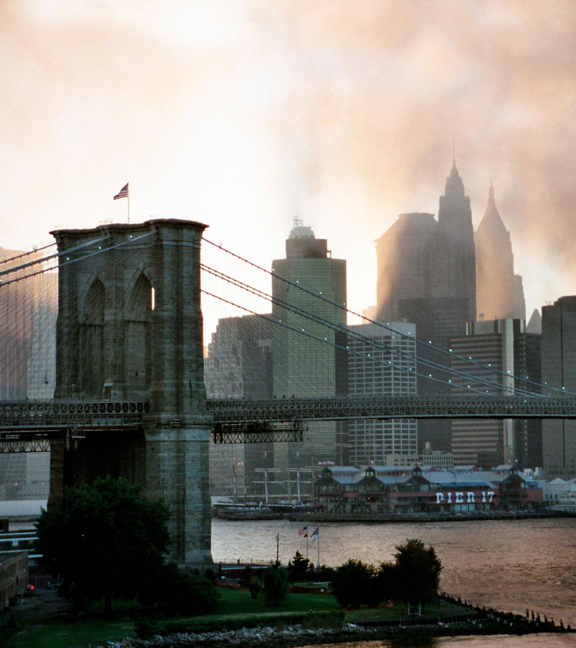 "ZDFzoom: 20 Jahre 9/11 - Ein Tag im September": Brooklyn-Bridge im Vordergrund, im Hintergrund Skyline von Manhattan im Sonnenuntergang und umhüllt von Rauch.