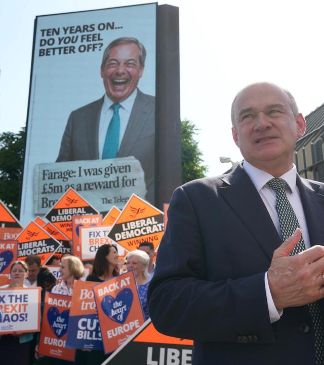 Der Vorsitzende der Liberaldemokraten, Ed Davey, steht vor einem Bildschirm, auf dem der Vorsitzende von Reform UK, Nigel Farage, zu sehen ist.