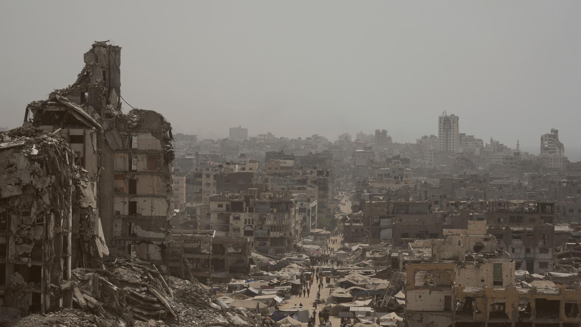 Menschen gehen eine Straße entlang, die von Gebäuden umgeben ist, die durch israelische Bombardierungen im Gazastreifen zerstört wurden.