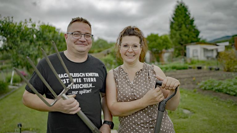 Eine Frau mittleren Alters lacht in die Kamera, in ihrer Hand hält sie einen Spaten. Neben ihr steht ein Mann mittleren Alters, der ebenfalls in die Kamera lacht. Er hält eine Mistgabel in der Hand. Im Hintergrund ist verschwommene Natur zu sehen.