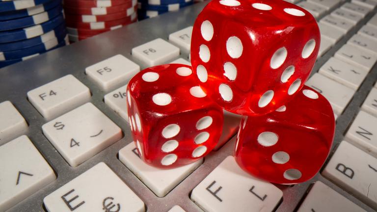 Symbolbild: rote Spielwürfel liegen auf der Tastatur eines Laptops. Daneben liegen bunte Spielchips. 