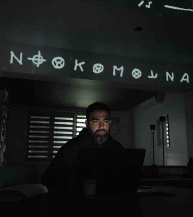 Ein Mann mit Bart sitzt in einem dunklen Zimmer, sein Gesicht wird durch den aufgeklappten Laptop vor ihm beleuchtet. Über ihm steht ein Schriftzug mit kryptischen Zeichen.