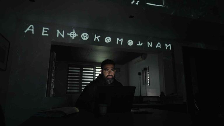 Ein Mann mit Bart sitzt in einem dunklen Zimmer, sein Gesicht wird durch den aufgeklappten Laptop vor ihm beleuchtet. Über ihm steht ein Schriftzug mit kryptischen Zeichen.