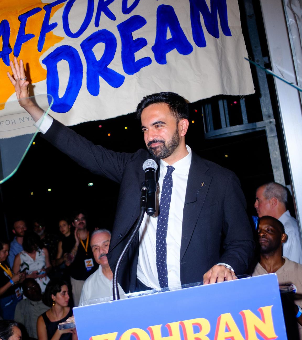 Zohran Mamdani steht an einem Rednerpult. Er hält einen Arm empor und lächelt dabei.