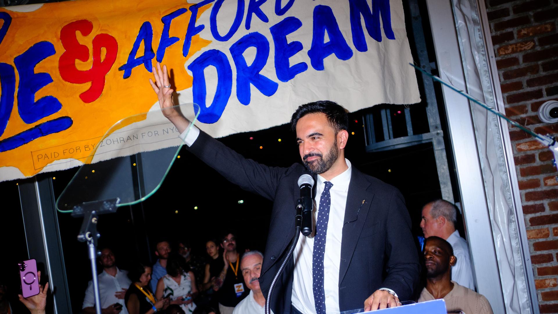 Zohran Mamdani steht an einem Rednerpult. Er hält einen Arm empor und lächelt dabei.