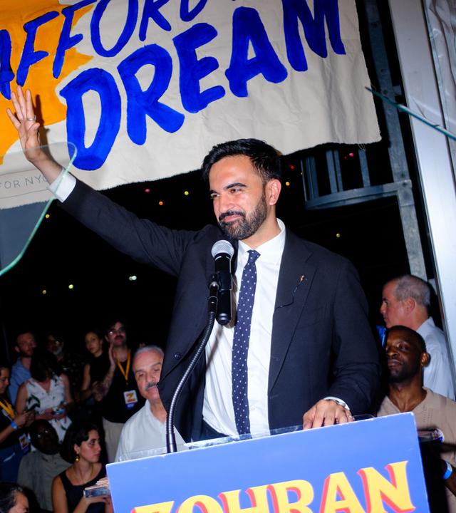 Zohran Mamdani steht an einem Rednerpult. Er hält einen Arm empor und lächelt dabei.
