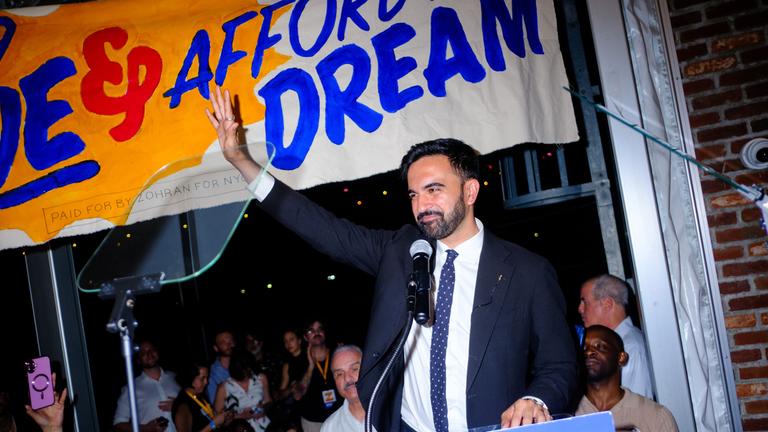 Zohran Mamdani steht an einem Rednerpult. Er hält einen Arm empor und lächelt dabei.