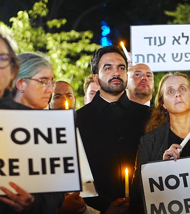 New Yorks Bürgermeister Zohran Mamdani auf der Mahnwache zum 7. Oktober in New York. Er schaut ernst an der Kamera vorbei, um ihn herum stehen Menschen mit Plakaten und Kerzen.