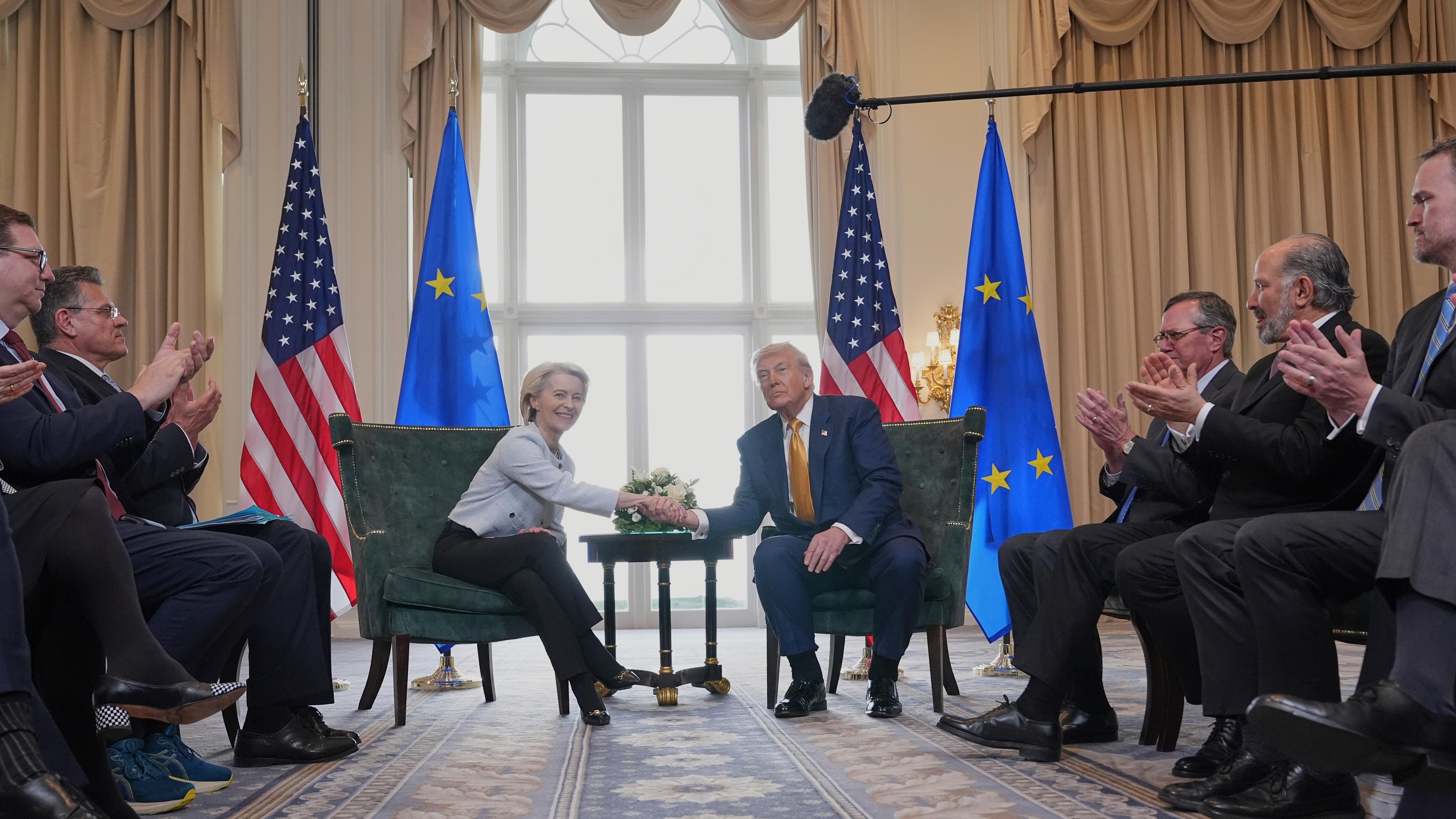 Ursula von der Leyen und Donald Trump geben sich zum Deal die Hände. Hinter ihnen stehen EU- und US-Flaggen