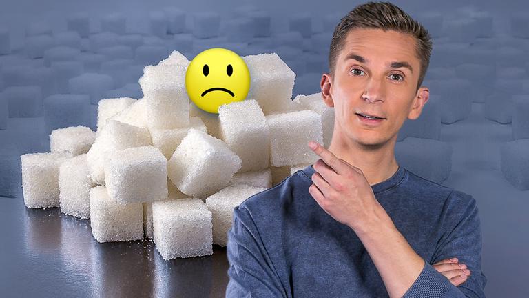 Eric Mayer im Vordergrund, dahinter jede Menge Würfelzucker. Eric zeigt auf einen kleinen Berg gestapelt aus Würfelzucker, auf dem ein negativer Smiley sitzt.