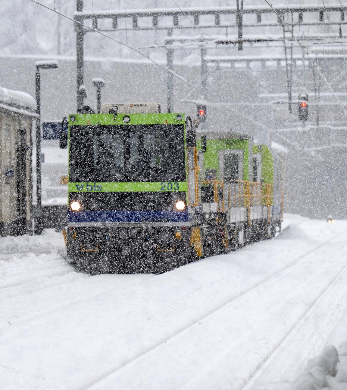 Bahnarbeiter schippen Schnee am Bahnhof Goppenstein. Es schneit und ein Hilfszug steht auf dem Gleis.