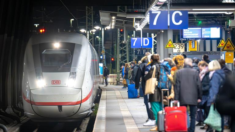 Zug fährt in Bahnhof ein und mehrere Menschen warten bereits am Gleis.