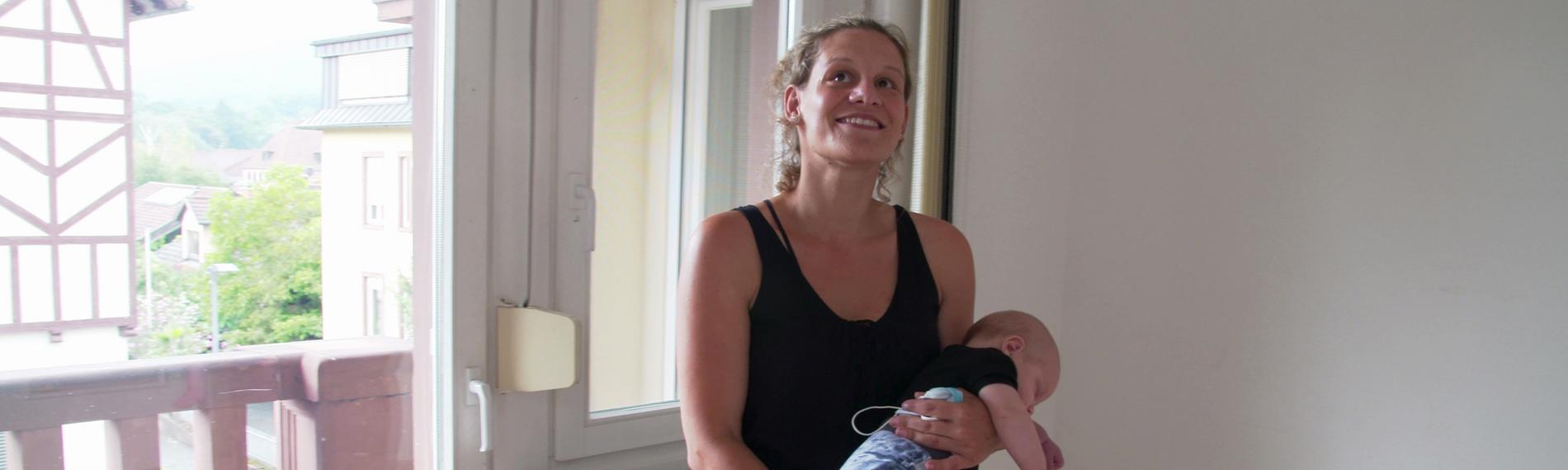 Eine junge Frau in einem leeren Raum vor der Balkontür stehend mit einem schlafenden Baby auf dem Arm. Sie lacht, wirkt hoffnungsvoll.