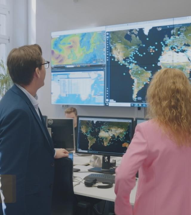 Eine Frau und ein Mann stehen in einem Büroraum. Sie blicken auf einen großen Bildschirm auf dem die Weltkarte gezeigt wird.