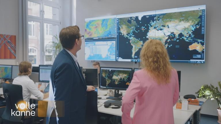Eine Frau und ein Mann stehen in einem Büroraum. Sie blicken auf einen großen Bildschirm auf dem die Weltkarte gezeigt wird.