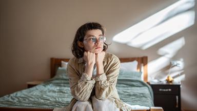 Junge Frau sitzt auf Bett und blickt Richtung Fenster, durch das Sonnenstrahlen in den Raum fallen.