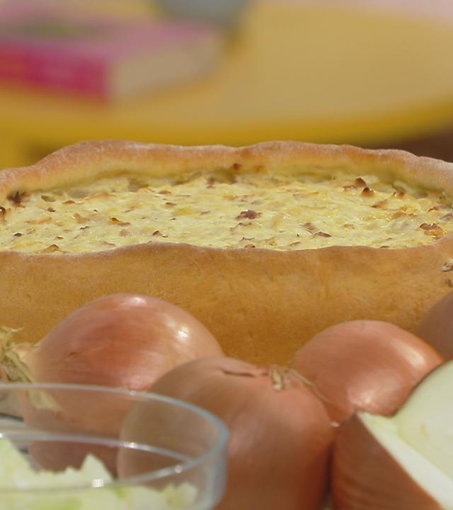 Zwiebelkuchen auf einem Tisch zu sehen, davor Zwiebeln.