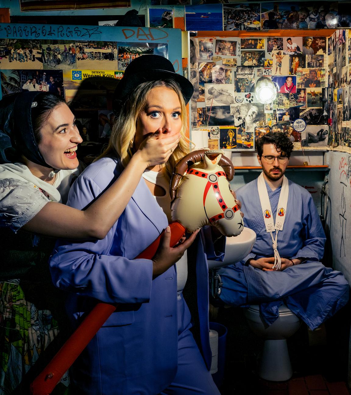 Atheistin Josi hält ein Steckenpferd in der Hand. Katholikin Gloria hält ihr dabei den Mund zu. Im Hintergrund meditiert Buddhist Darmasara auf der Toilette.